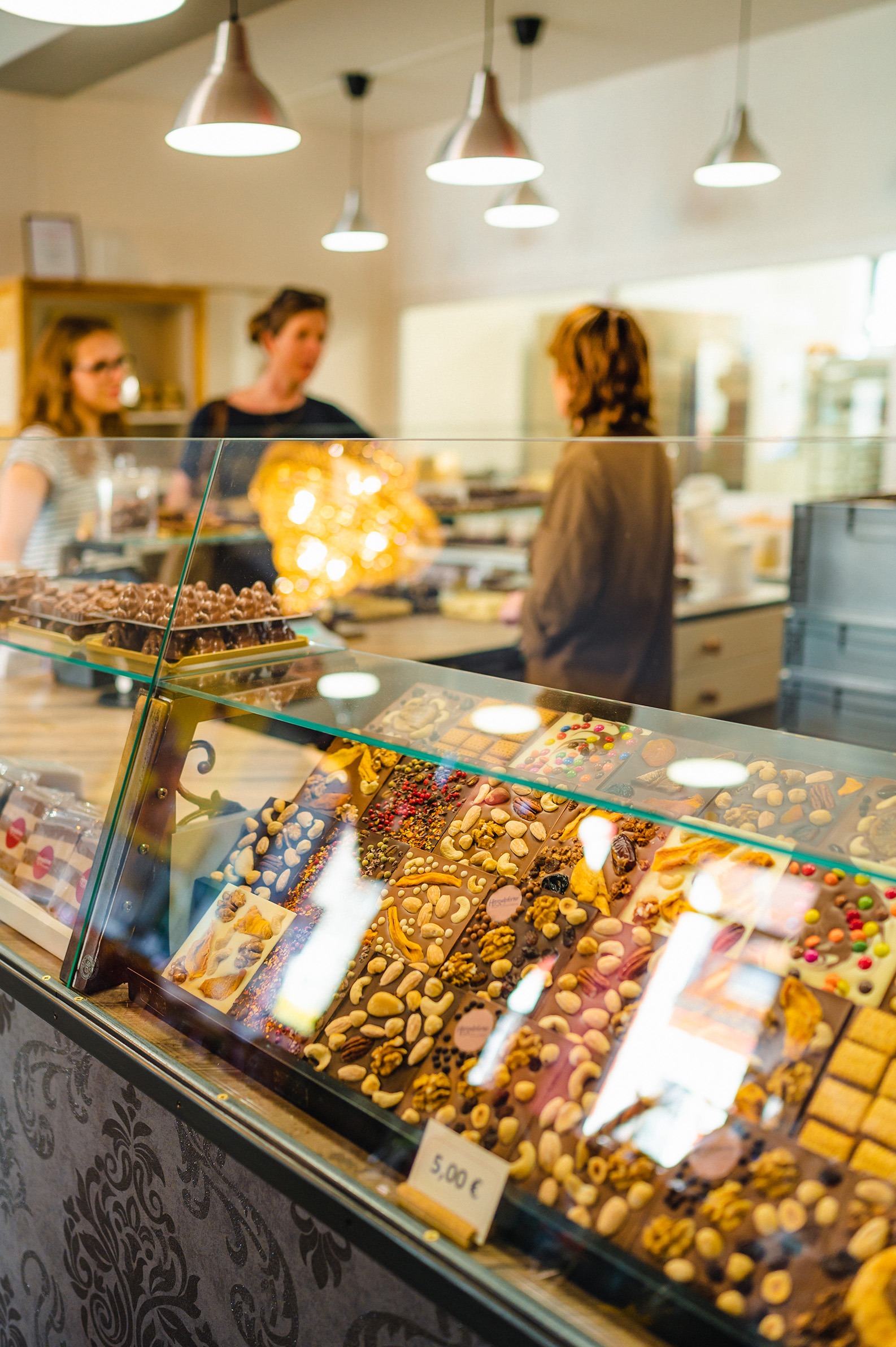 Nahaufnahme einer Theke mit einer vielfältigen Auswahl an Schokoladen; im Hintergrund zwei Frauen sich vom Personal beraten lassen.